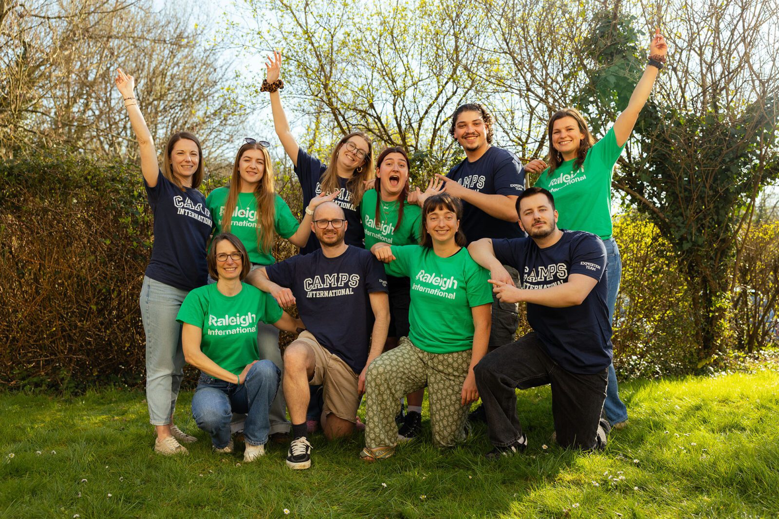Group of people wearing green Raleigh t-shirts and navy Camps International t-shirts posing for a photo outside with lots of greenery around them