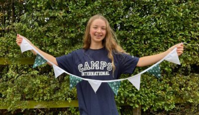 Teenage girl with long blond hair wearing a navy Camps International t-shirt and holding a handmade bunting with lots of greenery behind her