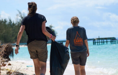 Two female student travellers on a Camps International expedition in Kenya, Africa. They're walking along Diani beach conducting a beach clean up of microplastics and washed up waste. They're wearing Camps International branded tshirts, walking away from the camera and carrying black bin bags full of rubbish.
