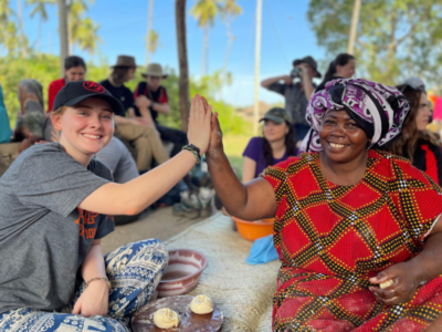 Girl traveller with a cap and grey t-shirt and a local woman from Tanzania dressed in traditional red patterned dress giving each other high five while making chapattis