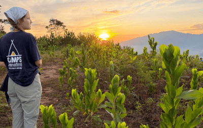 a student traveller girl wearing a camps t-shirt and a headband standing on a hill in Borneo and looking at the sunrise