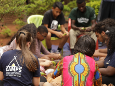 A group of students on a Camps International expedition sitting with a local group of African women, helping them to prepare food