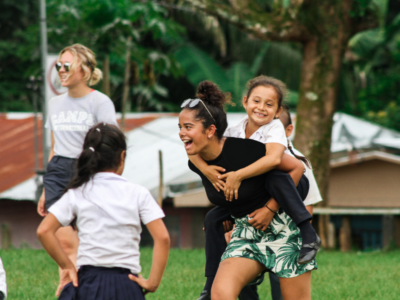 A couple of students on a Camps International expedition playing with some local school children. One student is smiling and carrying one of the children on her back, who's also smiling and laughing