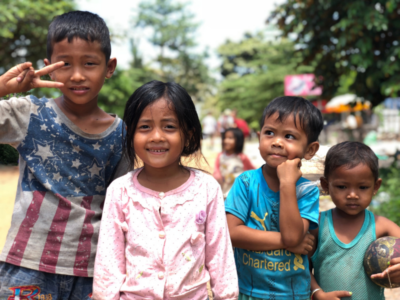 A group of four Cambodian children standing together, smiling at the camera and pulling faces