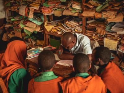 A group of African school children gathered around the desk of a teacher at school as he's writing in their books. The background has shelves full of paperwork and books, and the children are in their green and orange school uniforms