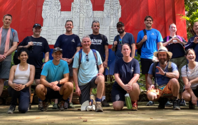 A group of teachers knelt on the floor in forest in front of the Cambodian flag, all smiling and happy on their familiarisation trip with Camps International